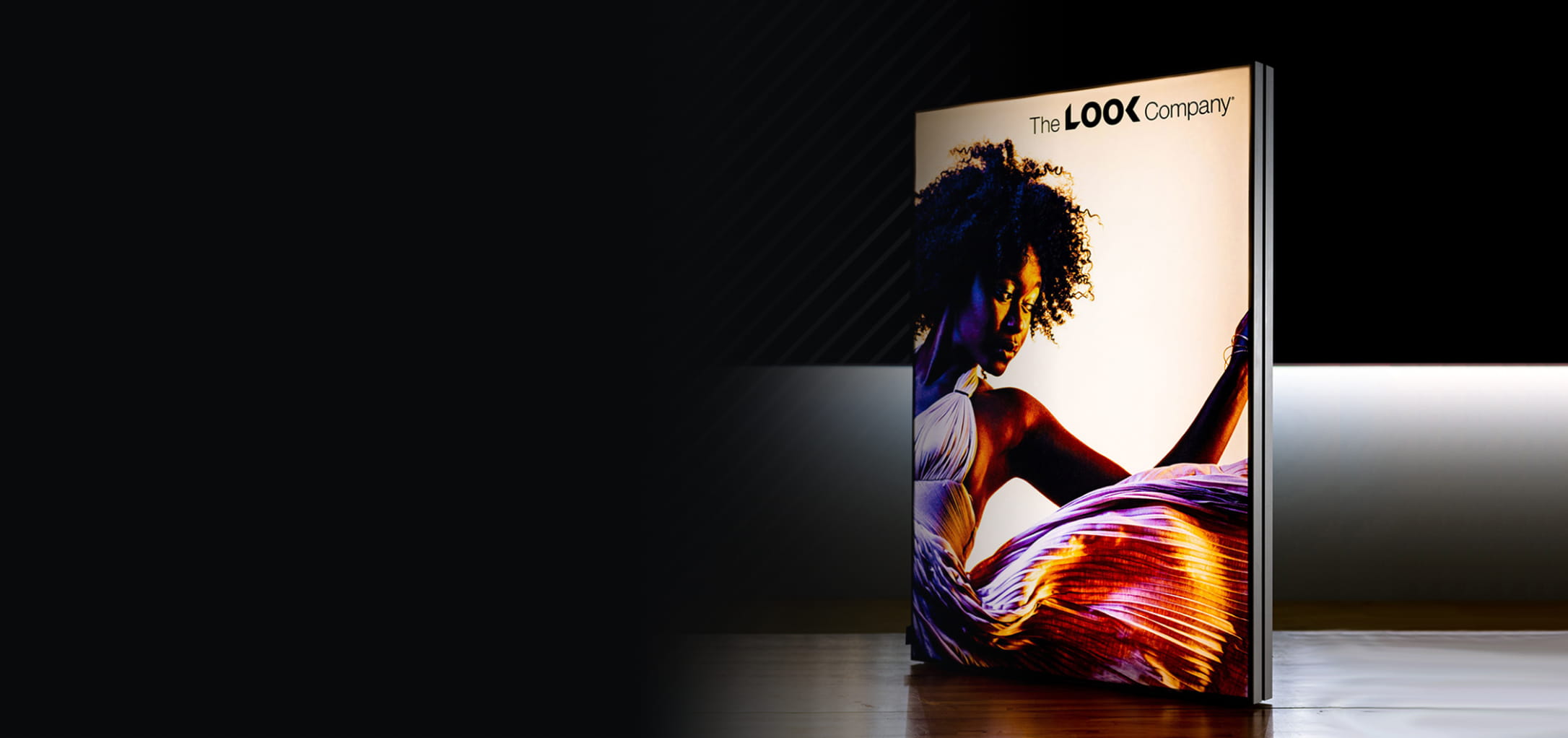 A slim, bright digital display features a colorful image of a woman and ""The LOOK Company"" logo, set on a reflective surface against a dark background.
