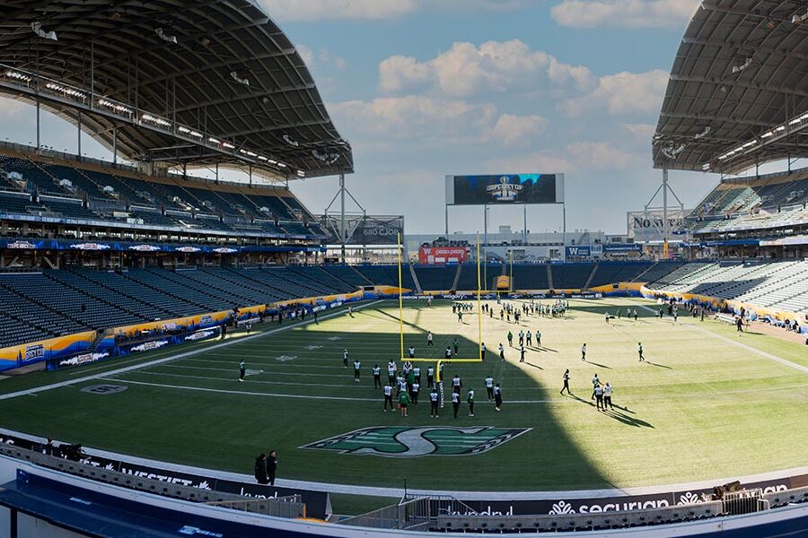 A wide shot of a modern football stadium with players practicing on the green field, mostly empty stands, and a Grey Cup logo on the scoreboard.