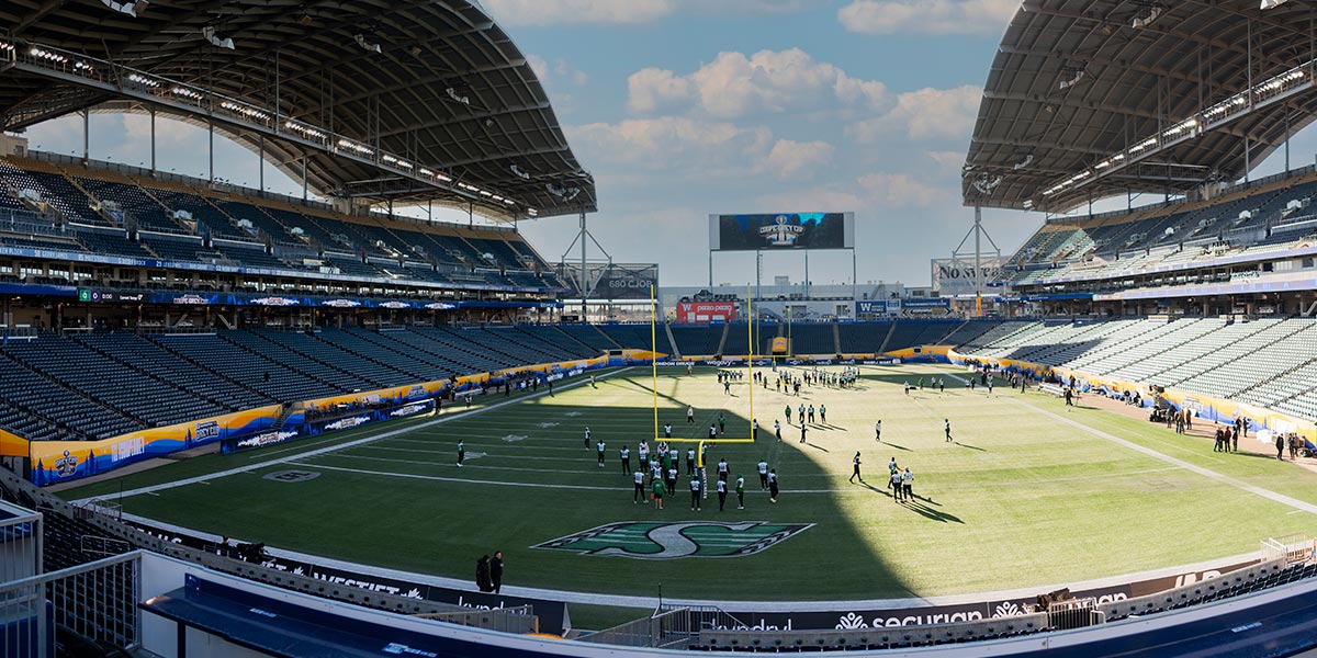 A wide shot of a modern football stadium with players practicing on the green field, mostly empty stands, and a Grey Cup logo on the scoreboard.