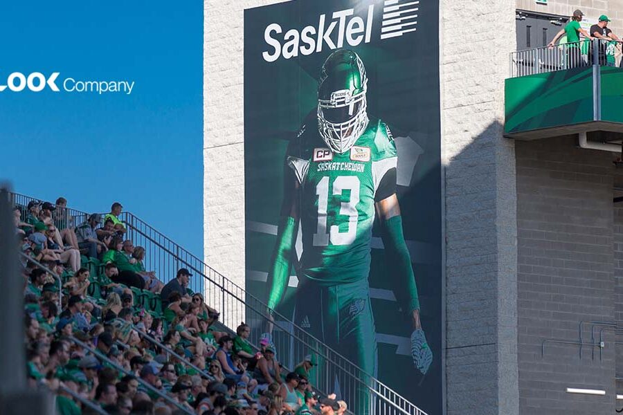 A stadium features a large banner of a green-uniformed football player, a crowd of fans in the stands, and ""The LOOK Company"" logo against a blue sky.