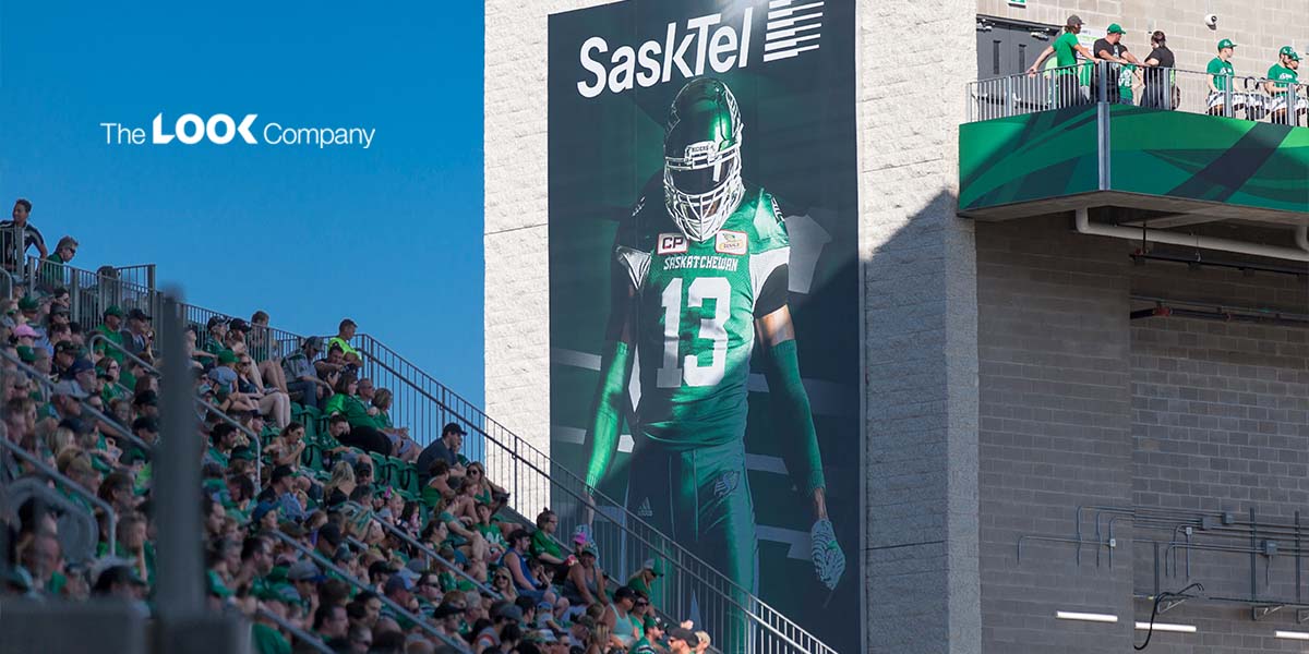 A stadium features a large banner of a green-uniformed football player, a crowd of fans in the stands, and ""The LOOK Company"" logo against a blue sky.