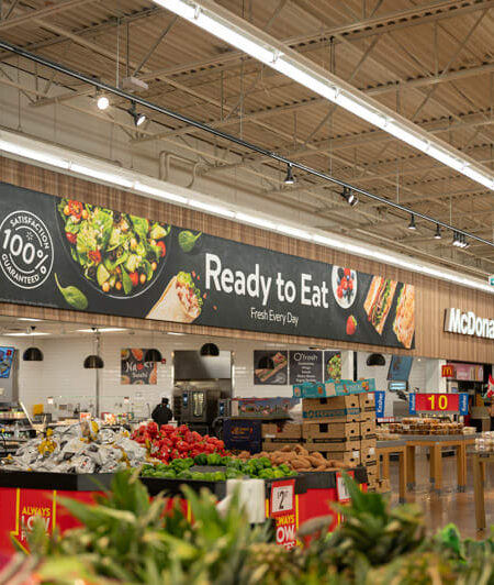 A brightly lit supermarket interior showcases a prominent ""Ready to Eat"" section with fresh produce displays, and a McDonald's restaurant in the background.