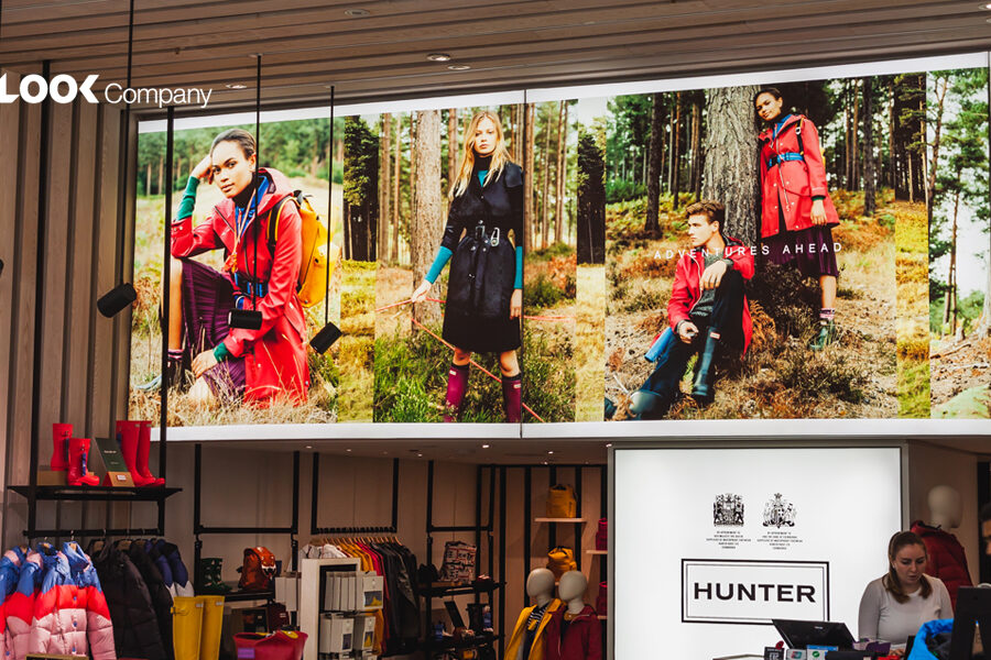 A retail store interior features a large banner with models wearing Hunter brand outdoor apparel and rain boots in a forest setting, alongside shelves displaying colorful boots and clothing racks.