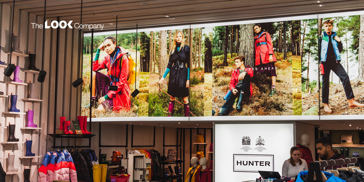 A retail store interior features a large banner with models wearing Hunter brand outdoor apparel and rain boots in a forest setting, alongside shelves displaying colorful boots and clothing racks.