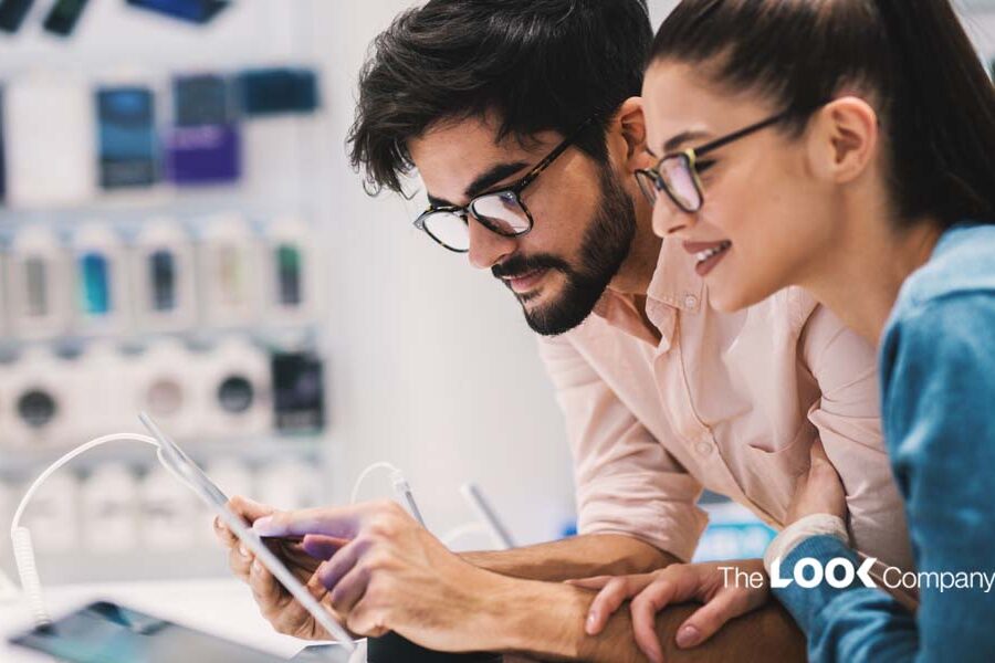 A man and woman wearing glasses look at a tablet together in an electronics store.