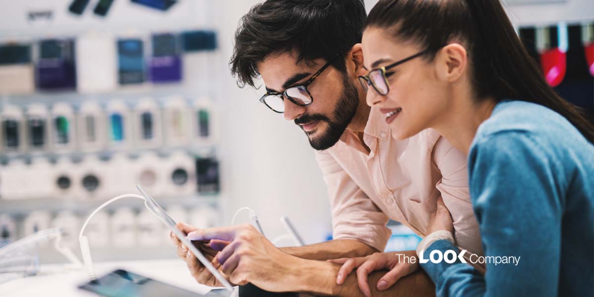 A man and woman wearing glasses look at a tablet together in an electronics store.