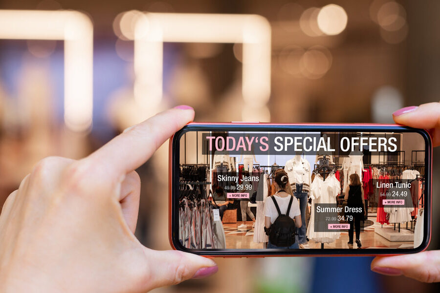 A smartphone held by hands displays augmented reality shopping offers overlaid on a clothing store scene.