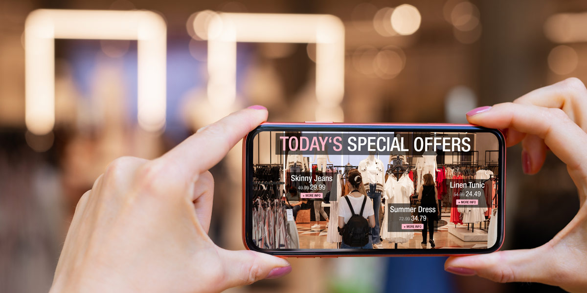 A smartphone held by hands displays augmented reality shopping offers overlaid on a clothing store scene. Demonstrating Omnichannel Retail Experience