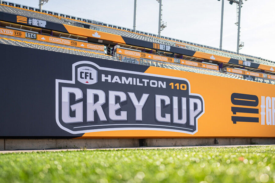 A large stadium banner advertises the ""CFL Hamilton 110 Grey Cup"" against a backdrop of stadium seating and green grass.
