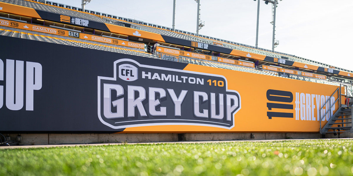 A large stadium banner advertises the ""CFL Hamilton 110 Grey Cup"" against a backdrop of stadium seating and green grass.