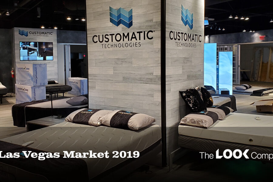 A modern Customatic Technologies showroom booth at the Las Vegas Market 2019, showcasing adjustable beds and mattresses.