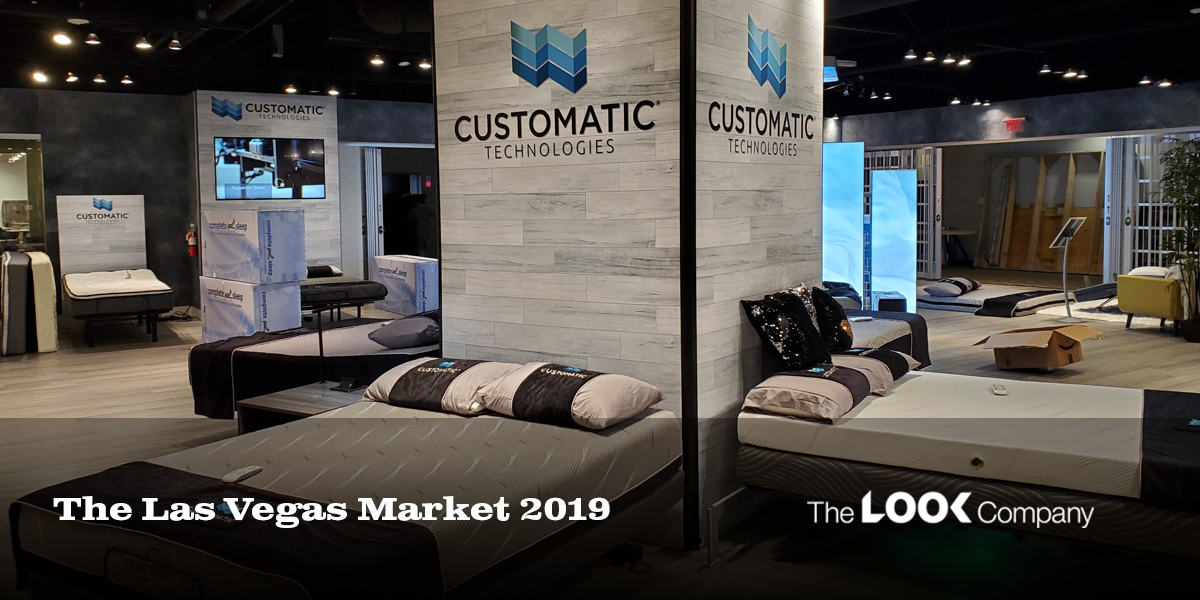 A modern Customatic Technologies showroom booth at the Las Vegas Market 2019, showcasing adjustable beds and mattresses.