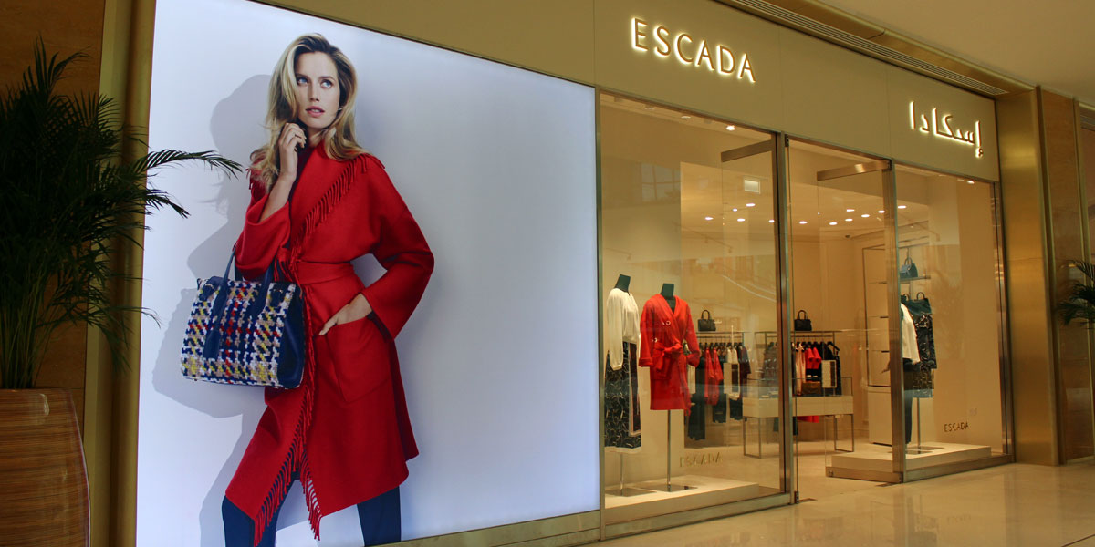 Escada store exterior featuring a large advertising poster of a model in a red coat and a patterned bag, next to its glass display window.
