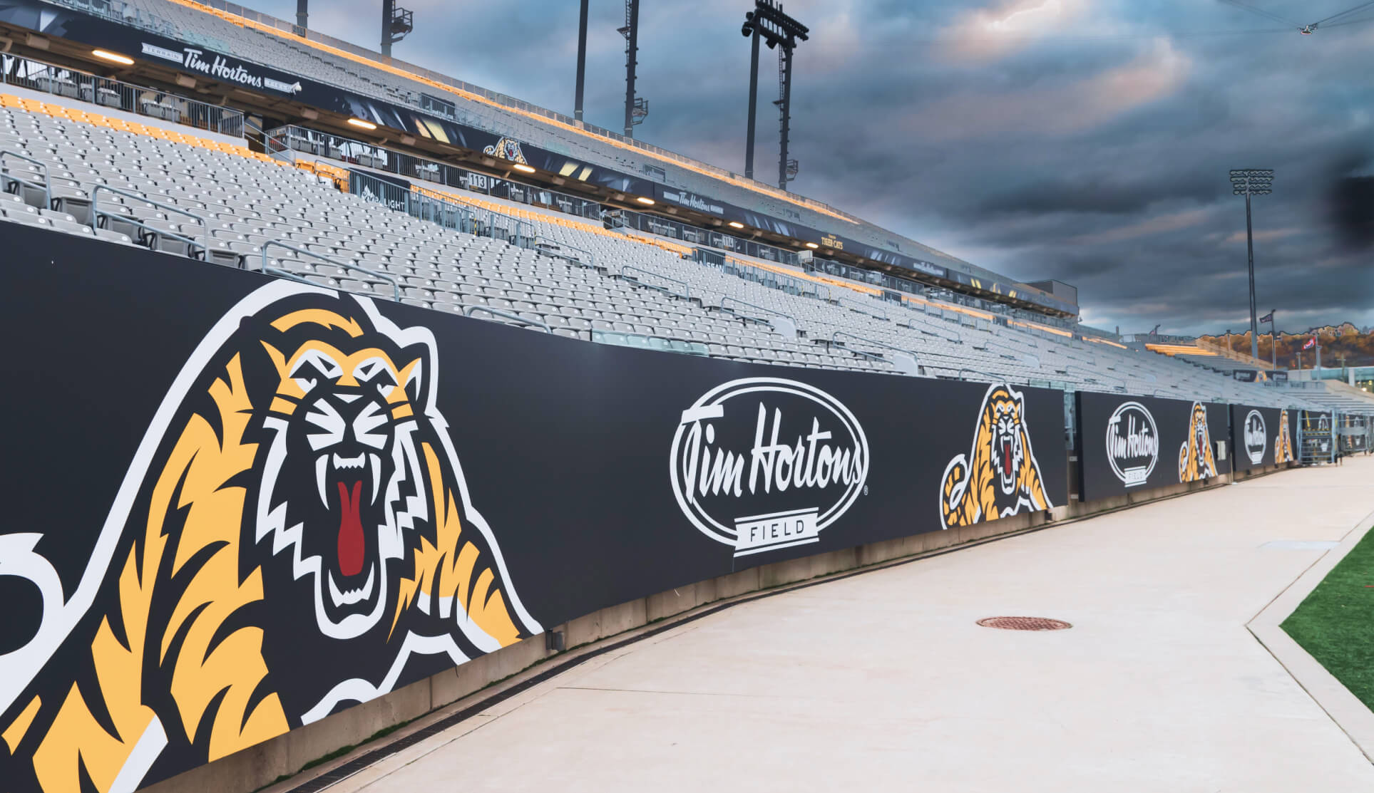 A low-angle shot of the sideline at Tim Hortons Field. The foreground features large black banners with a snarling yellow and black tiger logo and the Tim Hortons Field logo. In the background, empty grey stadium seating rises toward a cloudy, dramatic sunset sky.