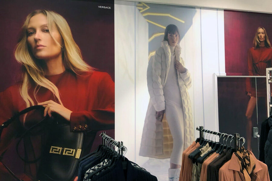 A clothing store interior features large fashion advertisements with models, including prominent Versace branding, and foregrounded clothing racks.