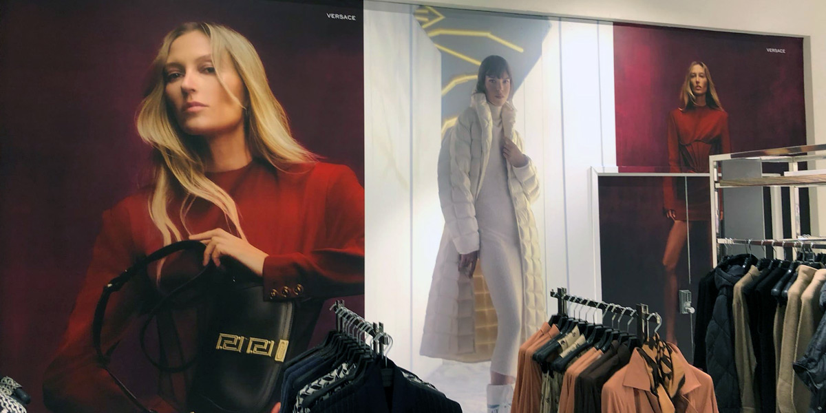 A clothing store interior features large fashion advertisements with models, including prominent Versace branding, and foregrounded clothing racks.