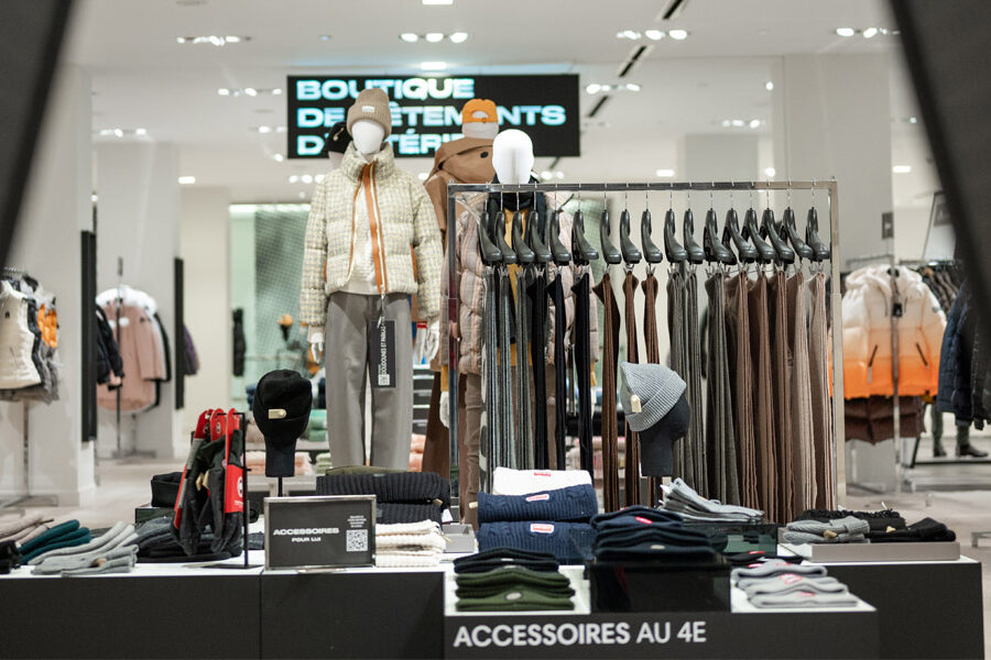 A bright clothing store interior features mannequins in winter wear, racks of apparel, and displays of hats and gloves.