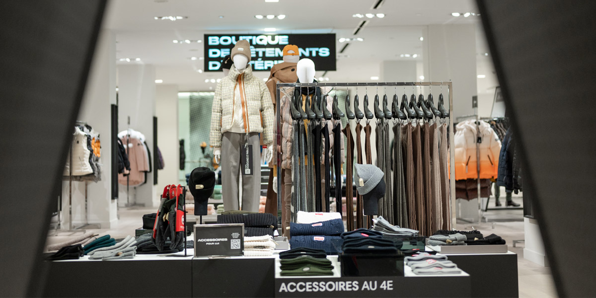 A bright clothing store interior features mannequins in winter wear, racks of apparel, and displays of hats and gloves.