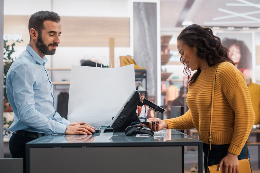 A woman makes a contactless payment with her phone at a retail store checkout counter attended by a male cashier.