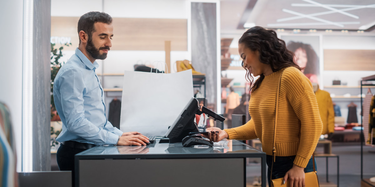 A woman makes a contactless payment with her phone at a retail store checkout counter attended by a male cashier.