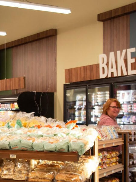 A brightly lit grocery store interior features a Bakery and Produce section, shelves stocked with various goods, refrigerated cases, and two people.