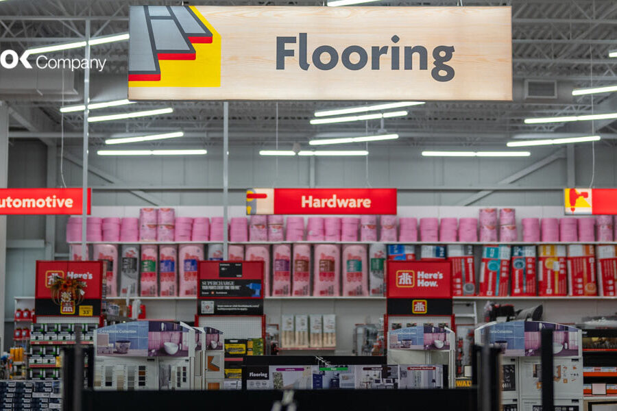 Interior of a brightly lit retail store featuring prominent overhead signage for Flooring, Automotive, Hardware, and Tools departments.