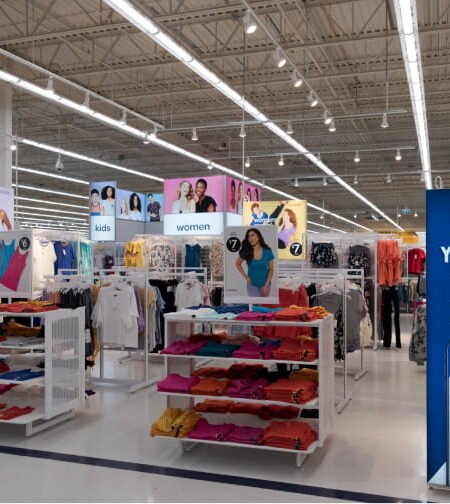 A brightly lit Walmart clothing department displays apparel on racks and tables, with overhead signs for ""kids"" and ""women,"" and a blue sign promoting online shopping via QR code.