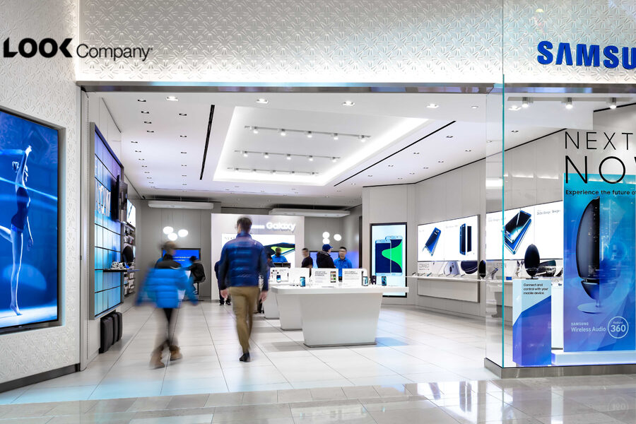 A modern Samsung retail store interior features numerous product displays, large video screens, and customers browsing.