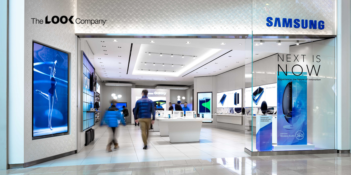 A modern Samsung retail store interior features numerous product displays, large video screens, and customers browsing.