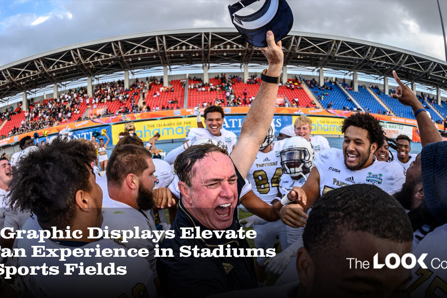 Excited football players and their coach celebrate on a field with a stadium full of fans and large graphic displays.