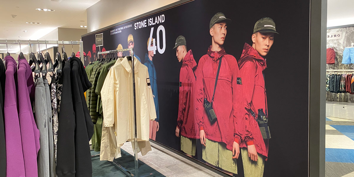 A clothing store display features racks of hoodies and jackets next to a large Stone Island 40 banner showing models wearing red coats and green shorts.