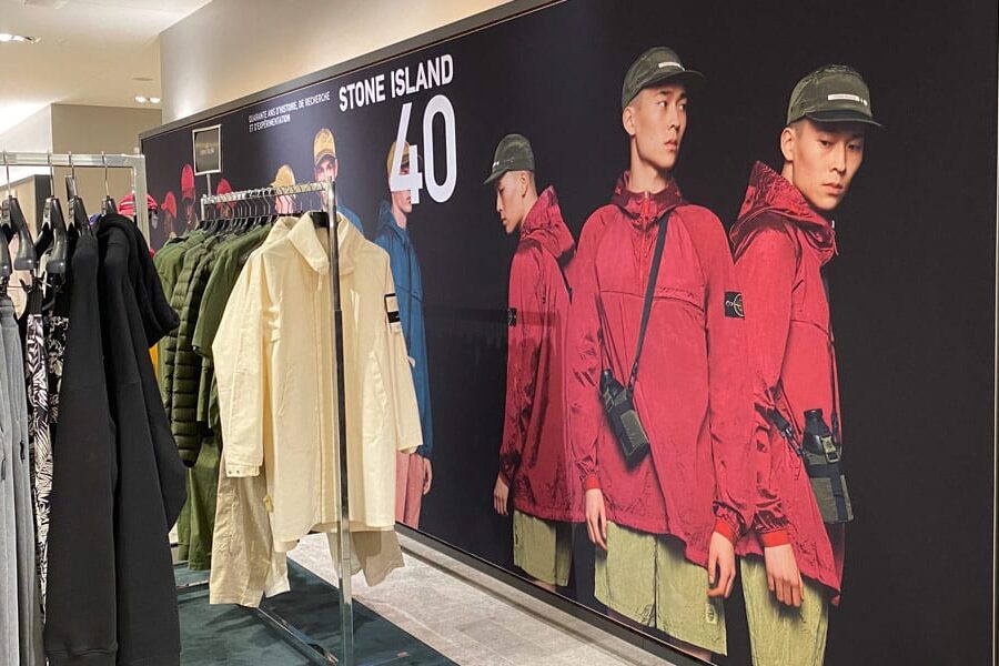A Stone Island retail display featuring models in red jackets on a large banner and racks of clothes in the foreground.