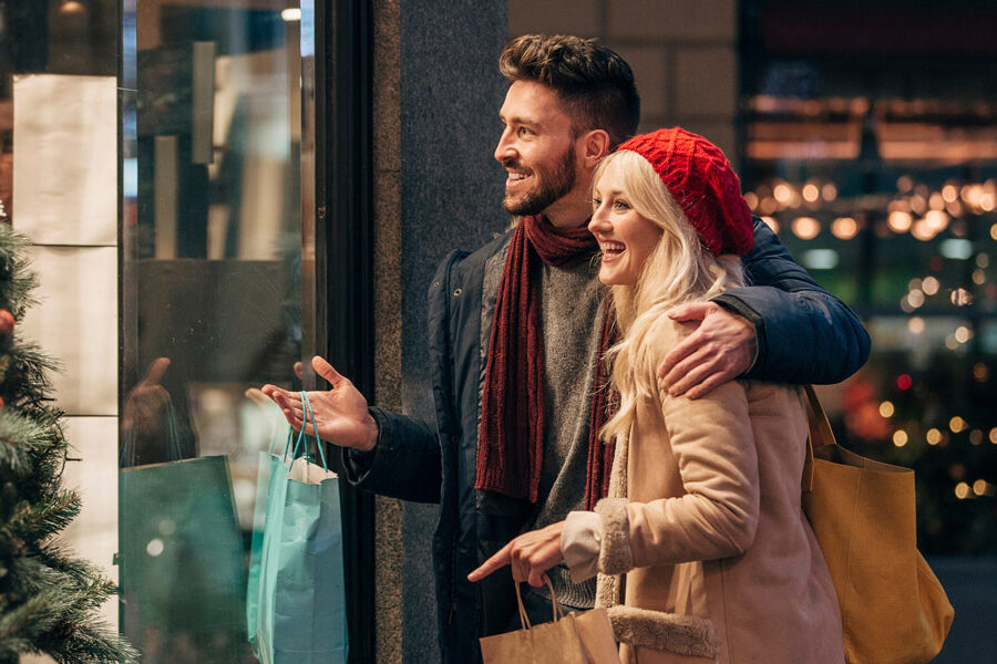 A happy couple smiles while looking into a festive store window with shopping bags.