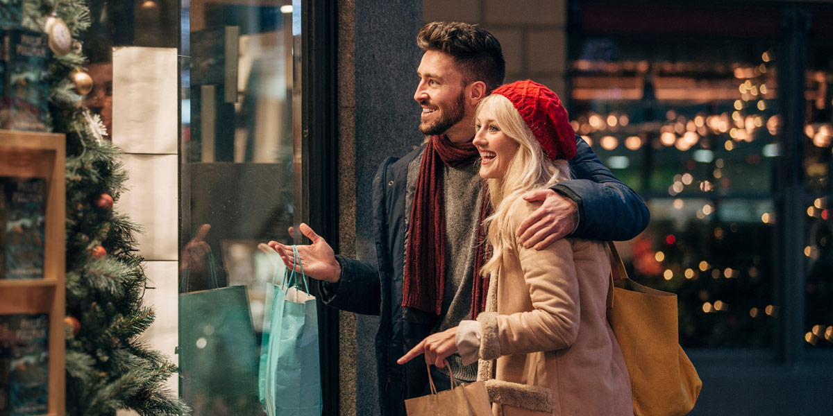 A happy couple smiles while looking into a festive store window with shopping bags.