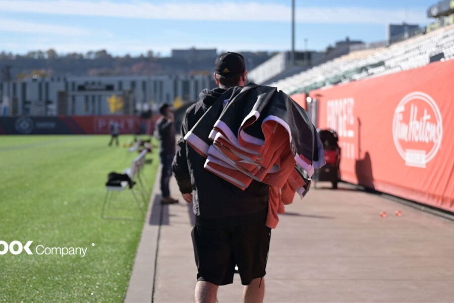 A person walks along a path next to a soccer field, carrying bundled items, with stadium stands and sponsor banners visible.