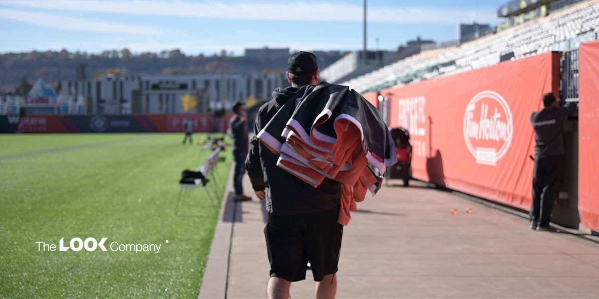 A person walks along a path next to a soccer field, carrying bundled items, with stadium stands and sponsor banners visible.
