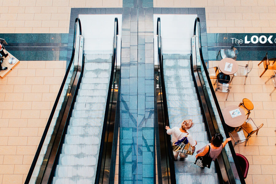 An overhead view of a modern shopping mall floor, showing two escalators, mannequins, cafe seating, and ""The LOOK Company"" logo.