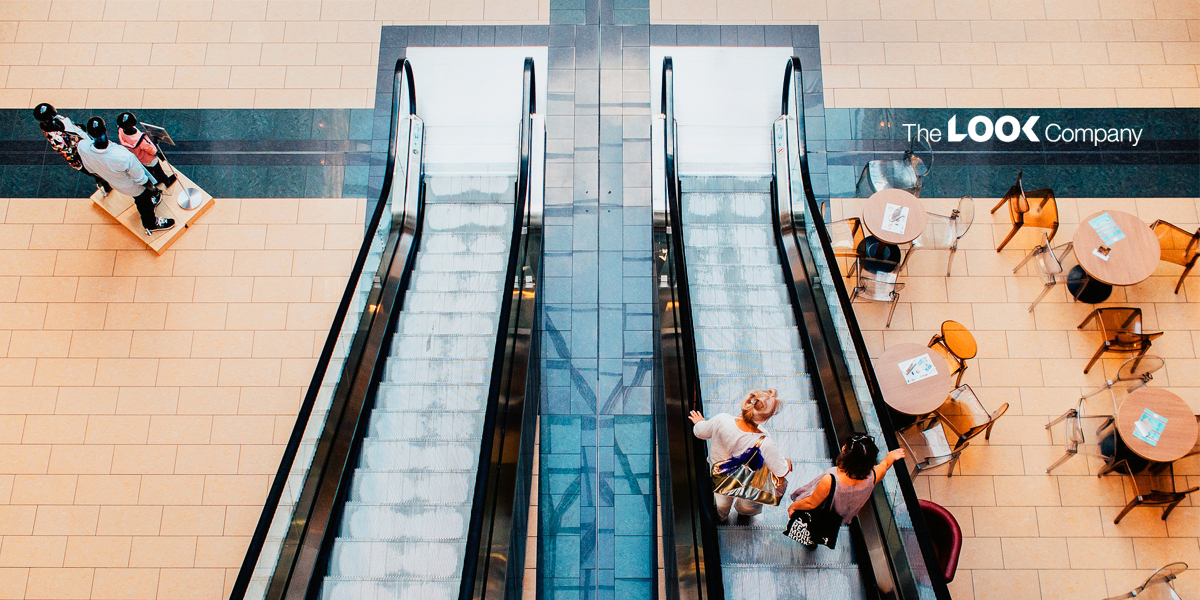 An overhead view of a modern shopping mall floor, showing two escalators, mannequins, cafe seating, and ""The LOOK Company"" logo.