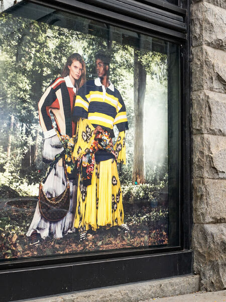 "A large shop window on a city street displays a ""The LOOK Company"" fashion advertisement featuring two models in colorful patterned outfits, with a tree trunk partially obscuring the left side."