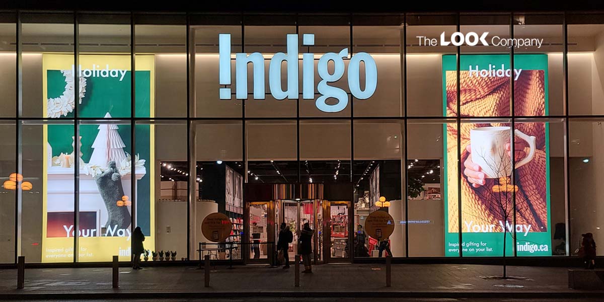Illuminated Indigo store facade at night with festive ""Holiday Your Way"" window displays.