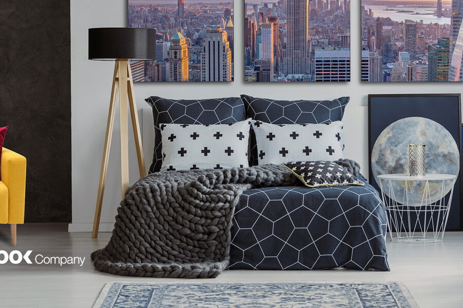 A modern bedroom features a bed with dark geometric bedding and a chunky gray blanket, complemented by cityscape wall art and a yellow sofa.