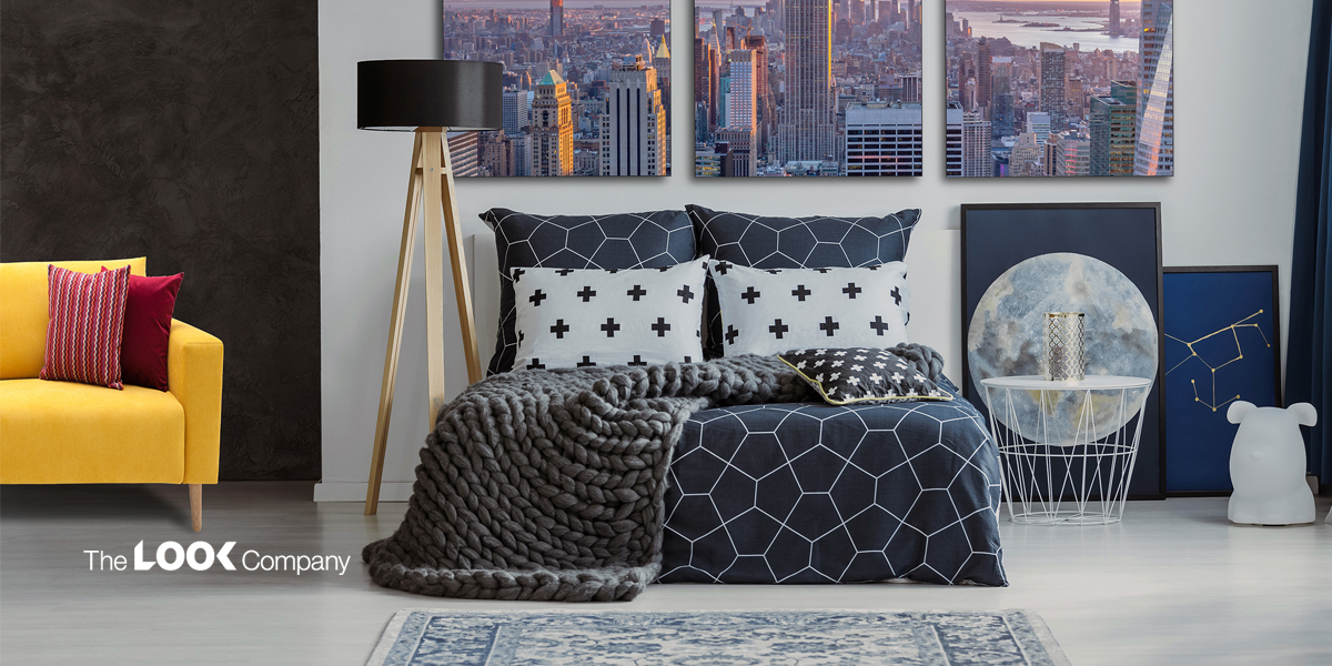 A modern bedroom features a bed with dark geometric bedding and a chunky gray blanket, complemented by cityscape wall art and a yellow sofa.