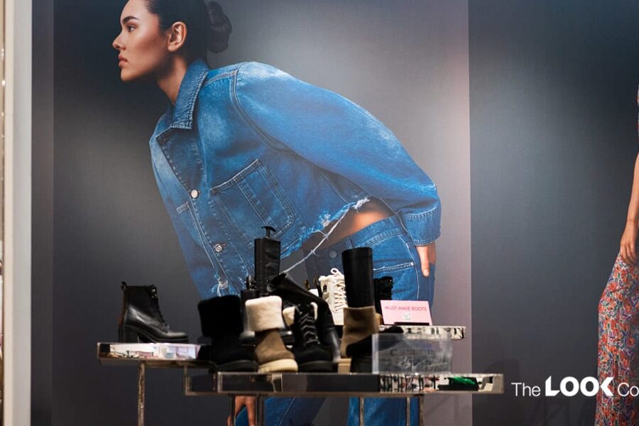 A retail store window features a display of boots in front of a large fashion advertisement showing models in denim and floral attire.