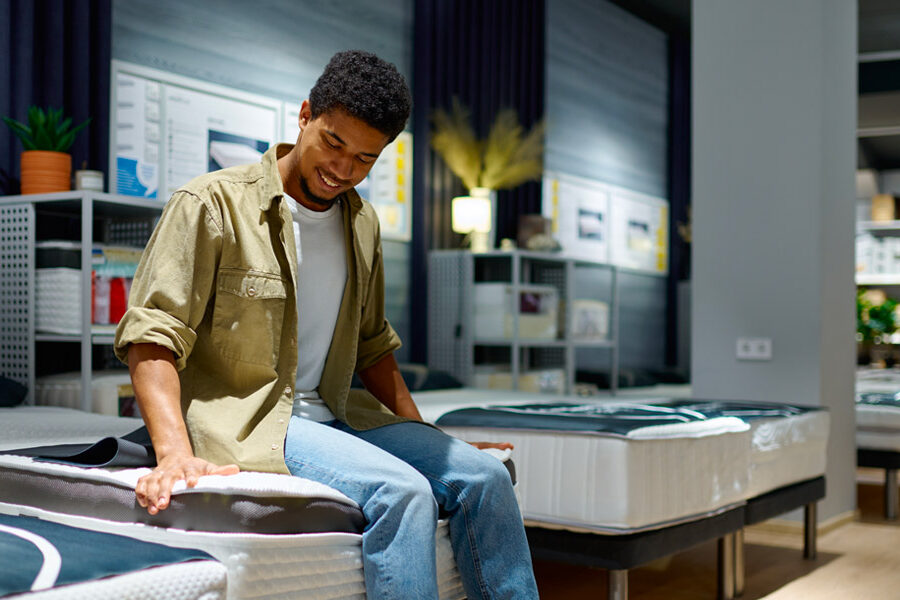 A smiling young man sits on and tests a mattress in a furniture store.