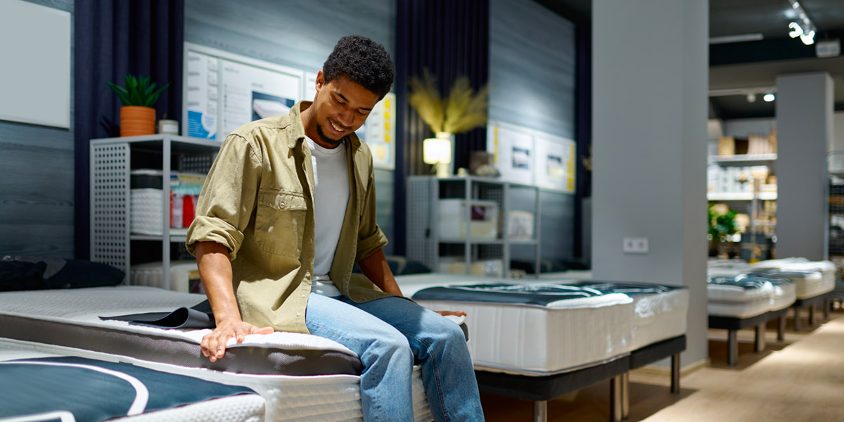 A smiling young man sits on and tests a mattress in a furniture store.