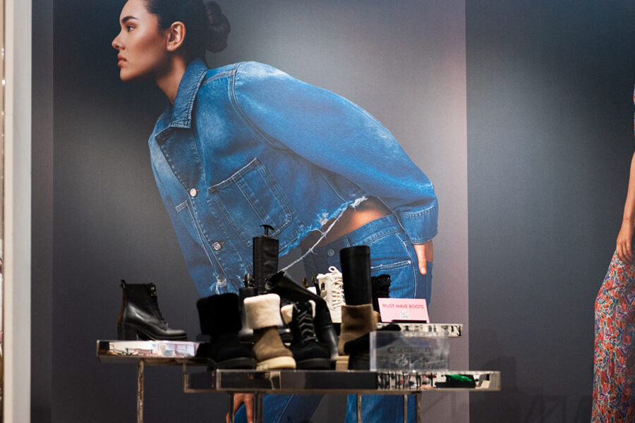 Store window display with large posters of women in a denim outfit and floral jumpsuit, showcasing various boots in the foreground.