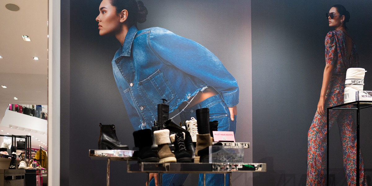 Store window display with large posters of women in a denim outfit and floral jumpsuit, showcasing various boots in the foreground.