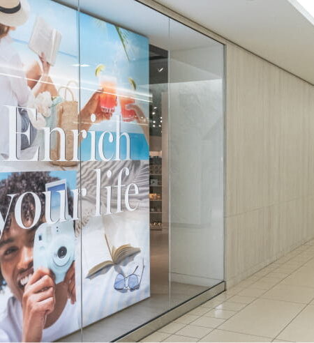A bright, modern hallway features a glass storefront displaying a lifestyle collage with the text ""Enrich your life,"" next to a long, empty corridor.