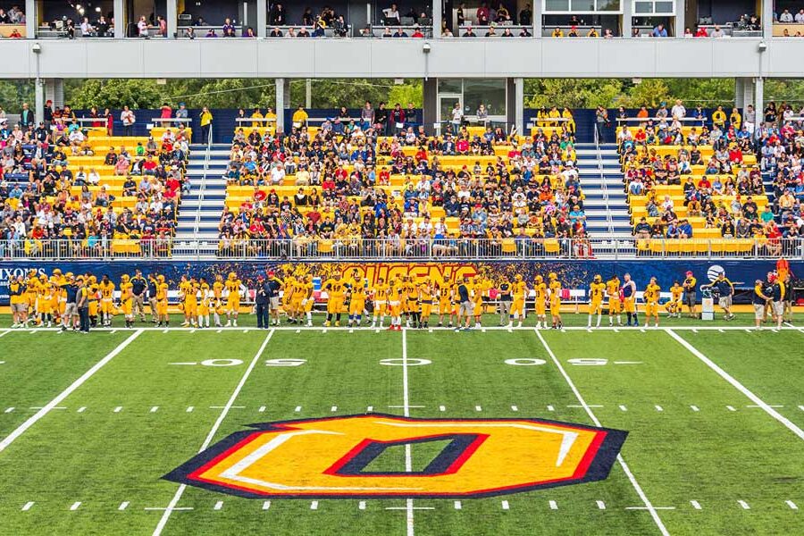 An American football team in yellow jerseys stands on a green field with a large ""O"" logo at midfield, overlooked by fans in stadium stands.