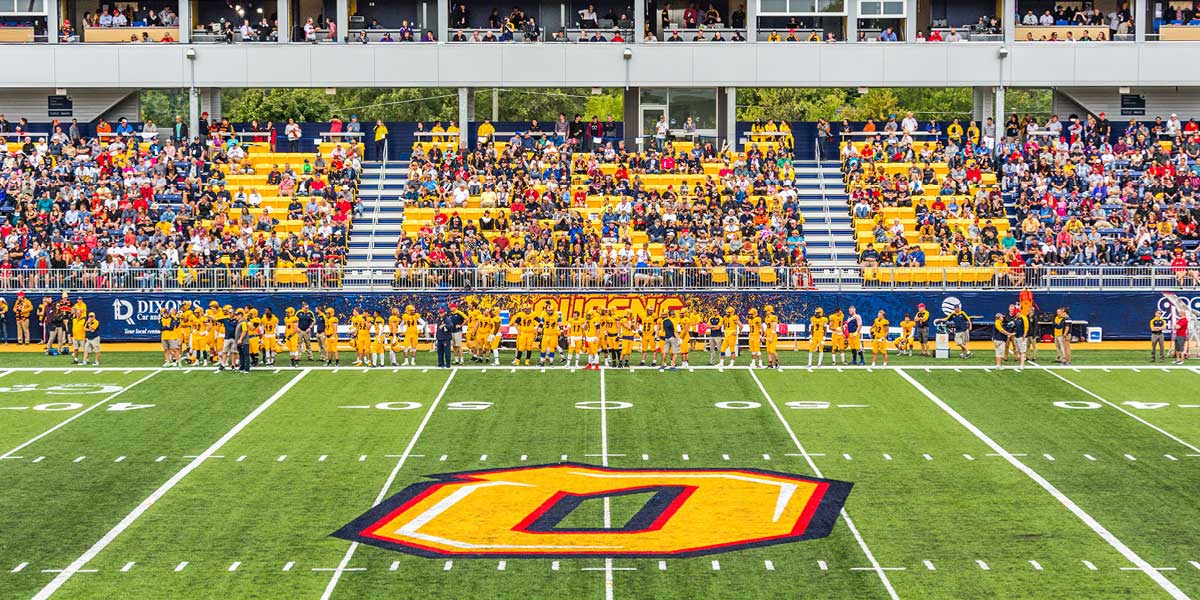 An American football team in yellow jerseys stands on a green field with a large ""O"" logo at midfield, overlooked by fans in stadium stands.